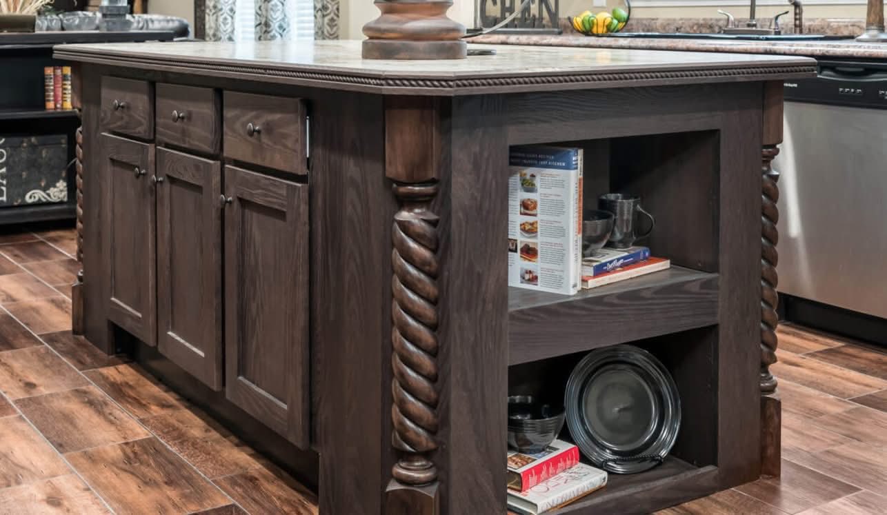 A dark wooden kitchen island with intricate spiral columns, shelves holding cookbooks and dishes, on a warm wood floor, conveying a rustic and cozy feel.