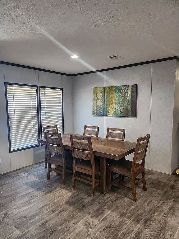 A cozy dining room with a wooden table and six matching chairs on a light wood floor. The wall art is abstract with green hues, next to two sunlit windows.
