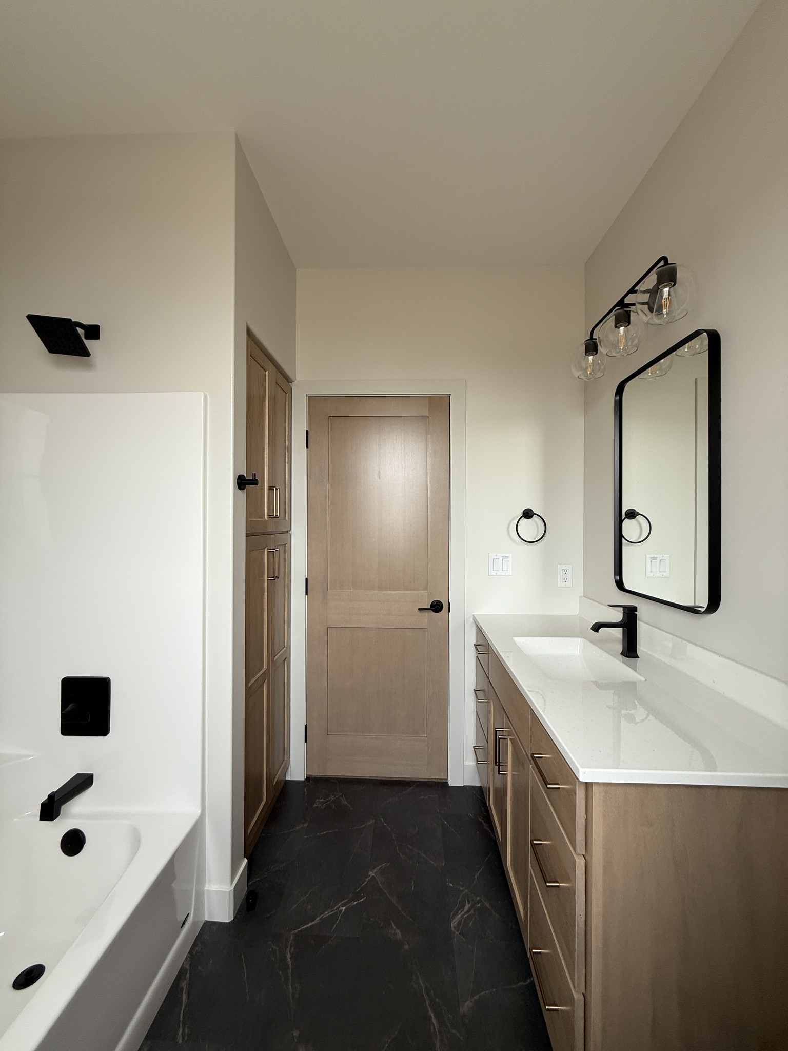 Sleek bathroom with a wooden vanity and white countertop. Black fixtures contrast with light walls. A tub is on the left, creating a modern, calm space.
