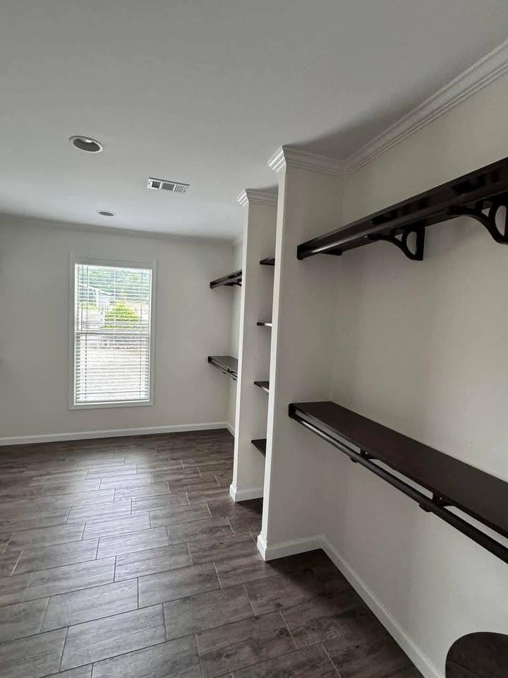 Minimalist walk-in closet with dark wooden shelves and rods on white walls, a single window with blinds, and dark wood flooring, creating an organized, modern feel.
