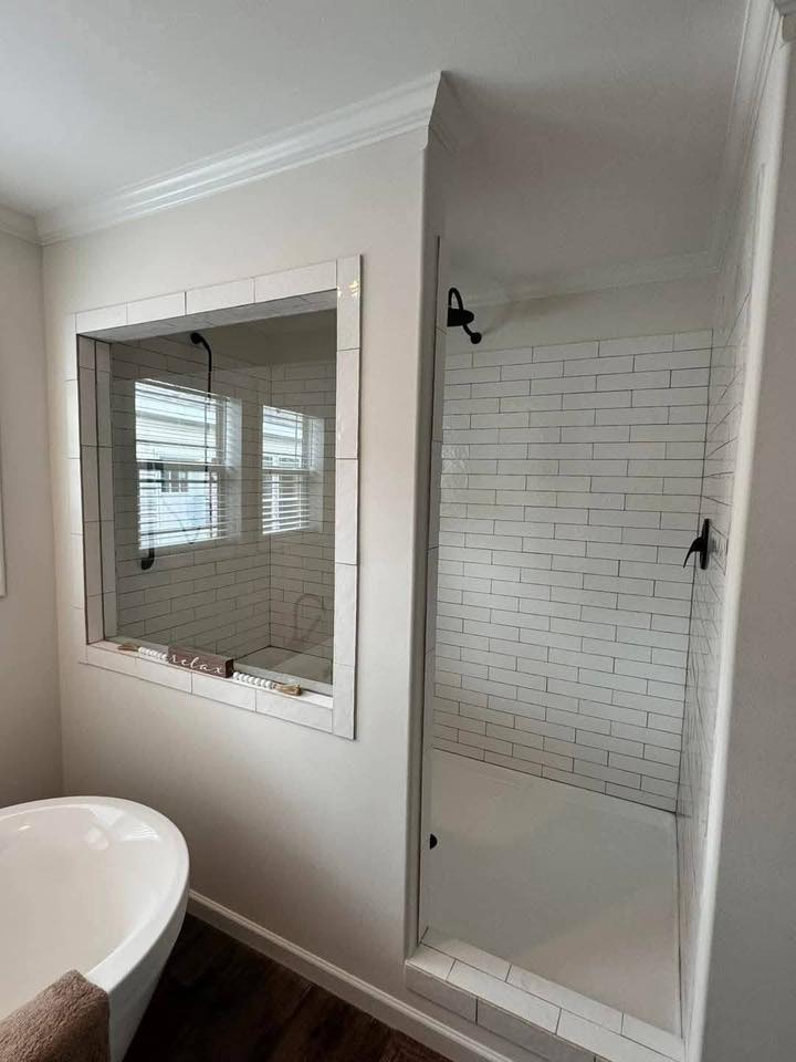 Modern bathroom with a white freestanding tub, a large window with blinds, and a tiled shower with glass door. The ambiance is clean and serene.