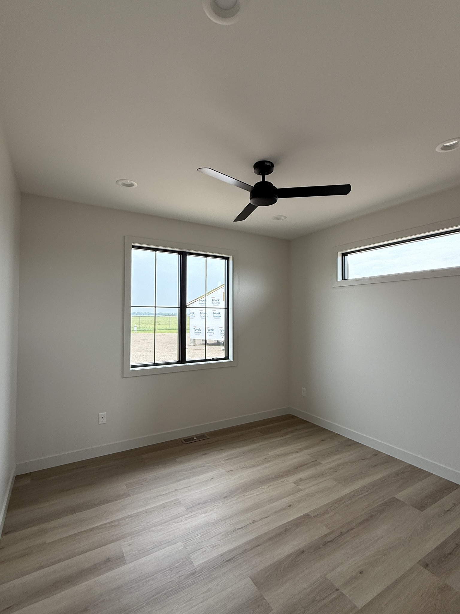 Minimalist room with light wood floors, white walls, a black ceiling fan, and two windows letting in natural light. Calm, serene atmosphere.