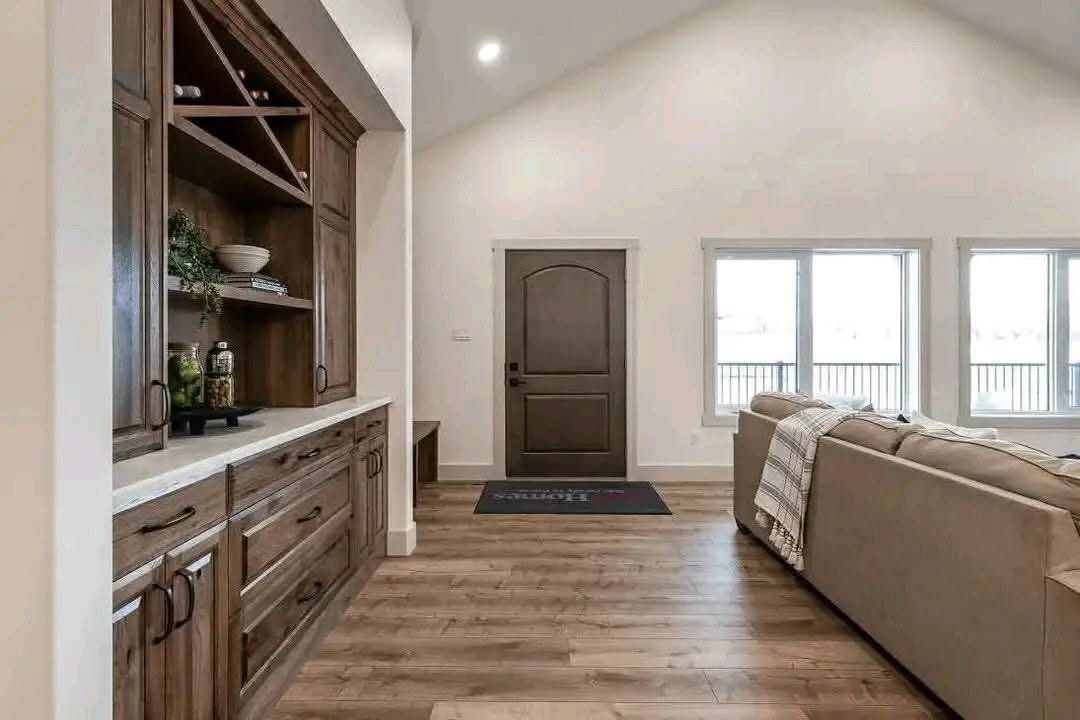 Spacious living room with wooden floors, a large tan sofa, and built-in wooden cabinets. Natural light streams through two windows, creating a warm ambiance.