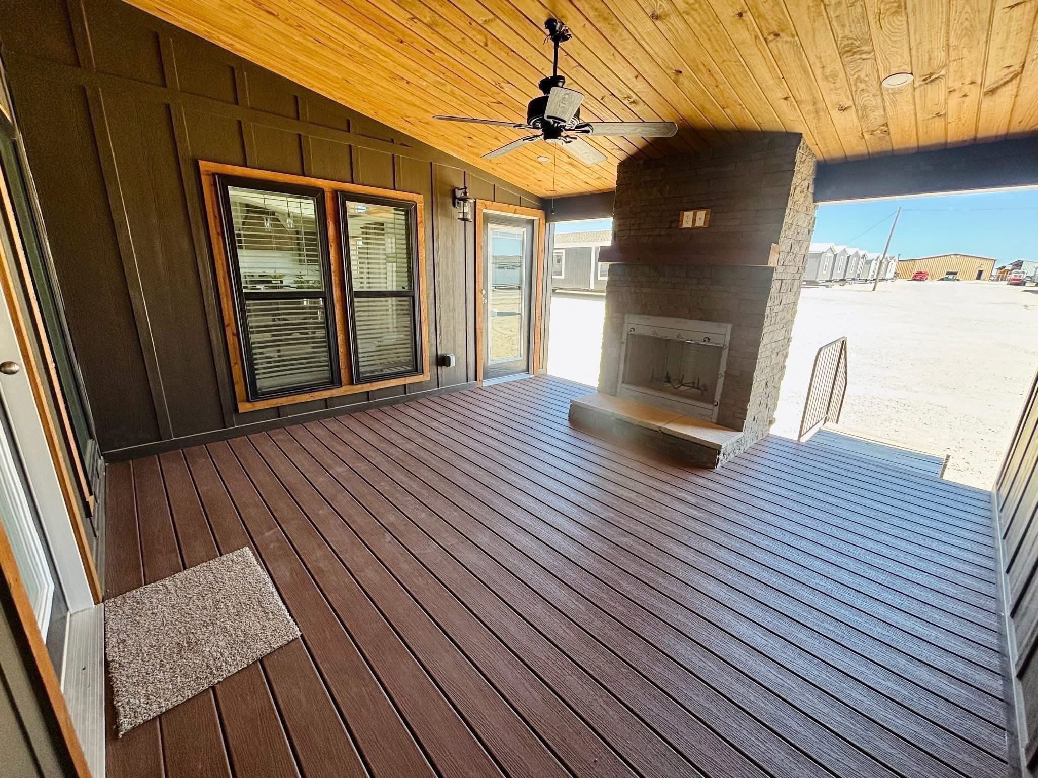 Covered porch with wooden deck and ceiling, brown siding, a brick fireplace, and ceiling fan. Sunlight streams through large windows, creating a cozy atmosphere.