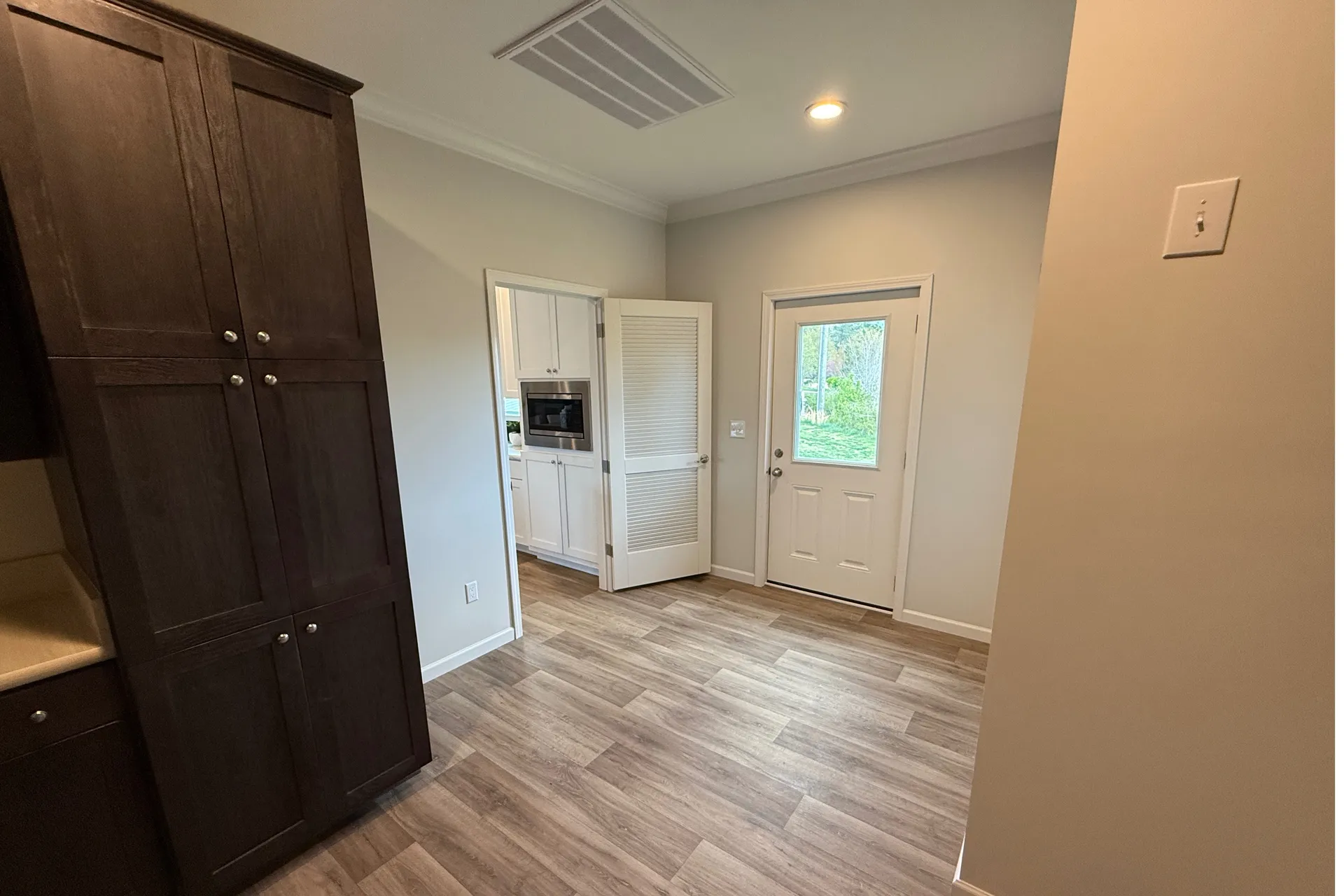 Spacious entryway with light wood flooring, dark wooden cabinets to the left, an open door leading to a white kitchen, and a glass door to the outside.