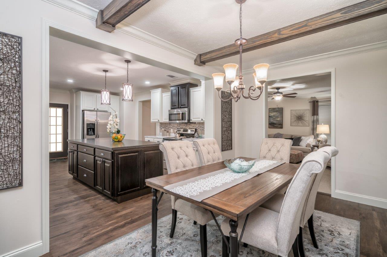 Elegant open-concept dining and kitchen area with wooden ceiling beams, a wooden table with tufted chairs, a modern chandelier, and a cozy living room visible.