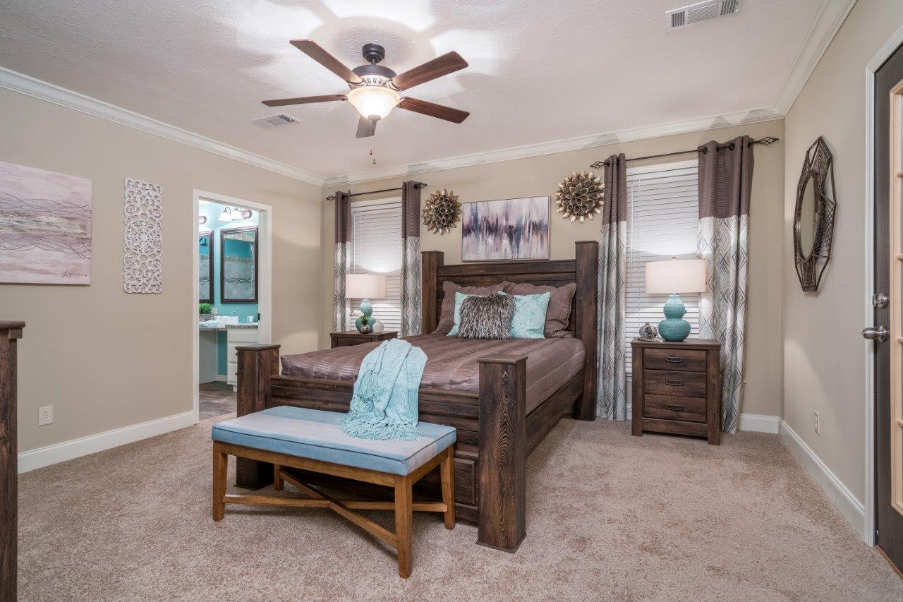 Cozy bedroom with a wooden bed, teal accents, and a ceiling fan. Two lamps on nightstands, art on walls, and an open door to a bathroom create a serene ambiance.