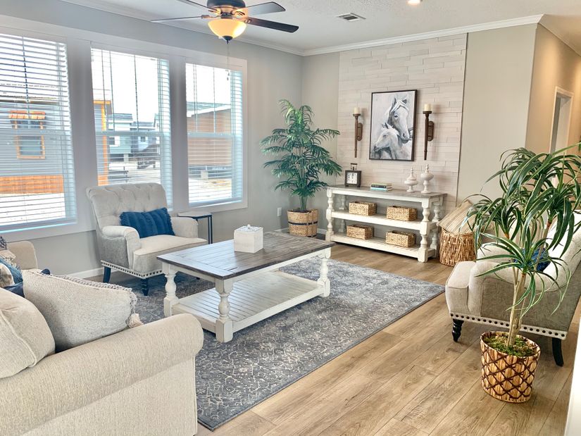 Bright, cozy living room with beige sofas and armchairs around a wooden coffee table. Large windows, indoor plants, and horse artwork create a serene vibe.