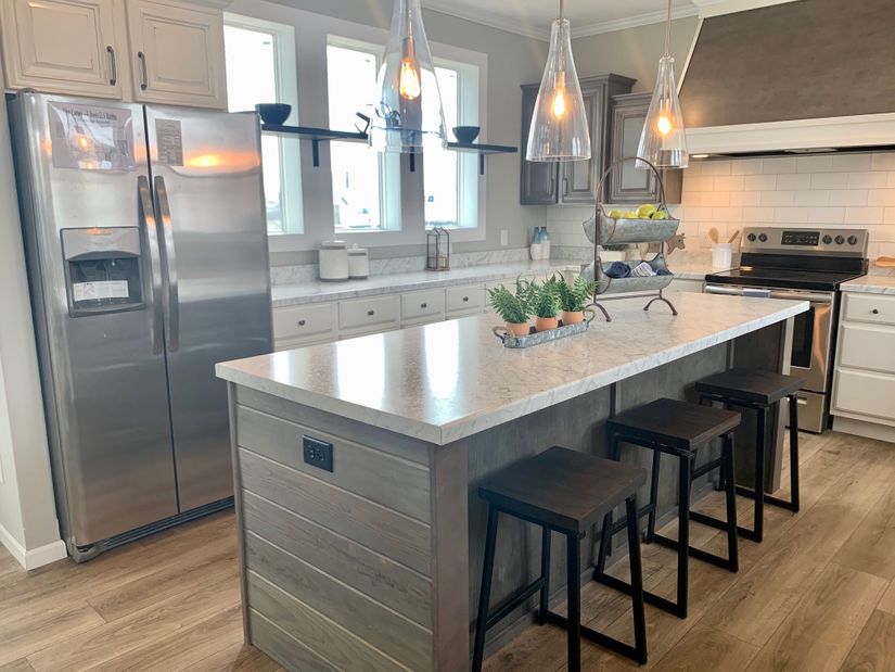 Modern kitchen with a gray island, three dark bar stools, and hanging pendant lights. Stainless steel fridge, potted plants, and bright windows.