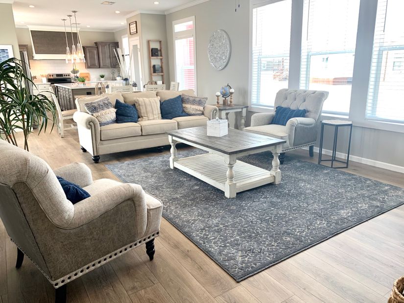 Spacious living room with beige sofas and navy cushions, a gray rug, and a central wooden coffee table. Bright natural light fills the room.