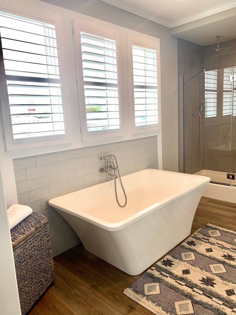 Bright bathroom featuring a freestanding white tub with chrome fixtures, wooden floors, and a patterned rug. Three shuttered windows add a fresh ambiance.