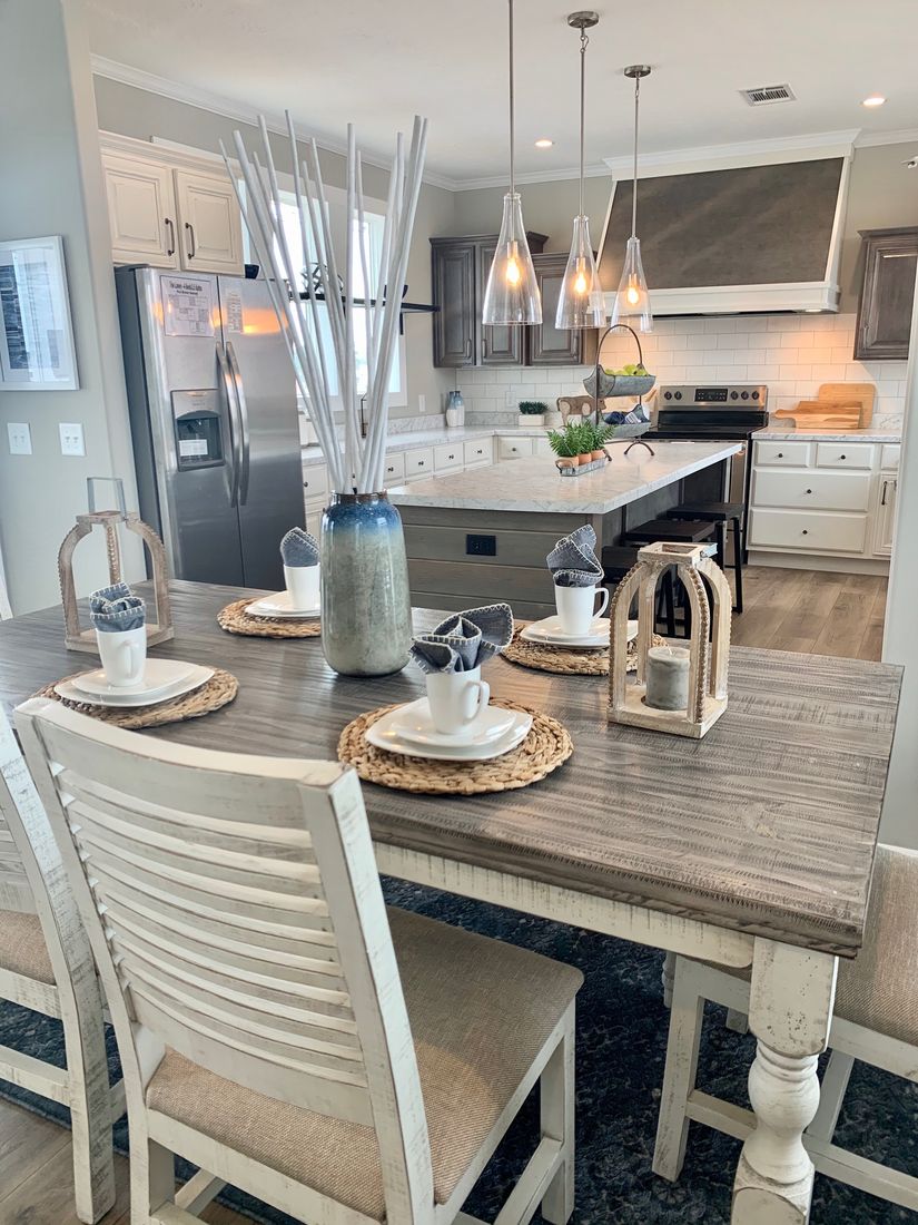 Rustic kitchen with a wood dining table set for four, featuring woven placemats, white cups, and lanterns. Modern pendant lights hang over a large island, with stainless steel appliances and white cabinetry in the background.