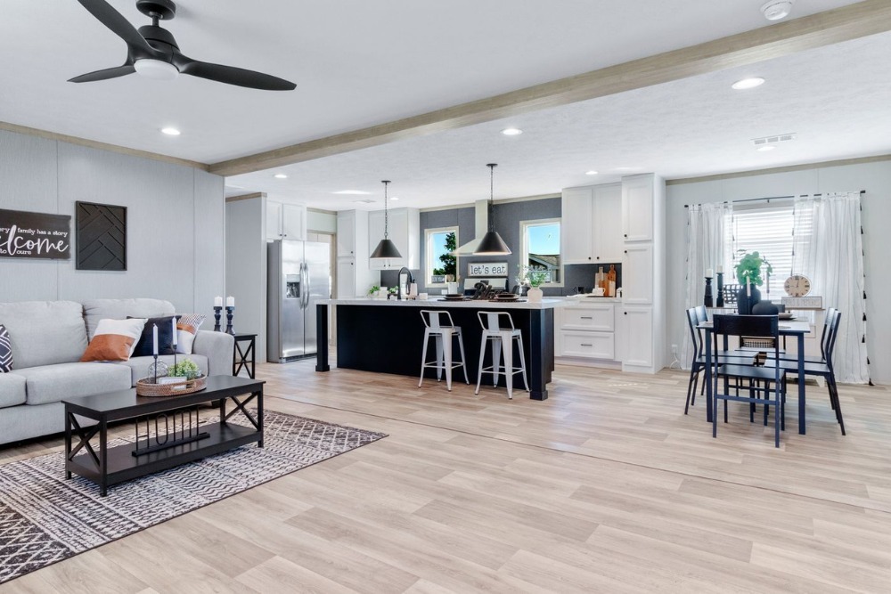 Modern open-plan living space with light wood flooring. Features a cozy beige sofa, stylish kitchen with bar stools, and a dining area under pendant lights.