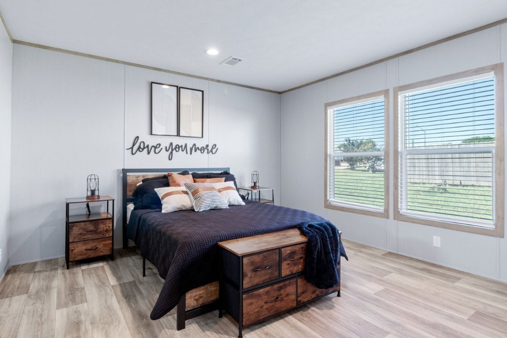 Modern bedroom with wood flooring, featuring a double bed with navy bedding and colorful pillows. "Love you more" sign above bed. Two large windows on the right and small bedside tables with lamps enhance the bright, cozy atmosphere.