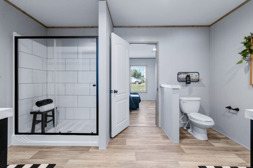 Modern bathroom with wooden floors, featuring a glass shower with a black frame, white tiles, and a stool. Adjacent is a toilet with decor. The open door reveals a glimpse of a blue room and outside greenery, creating a fresh ambiance.
