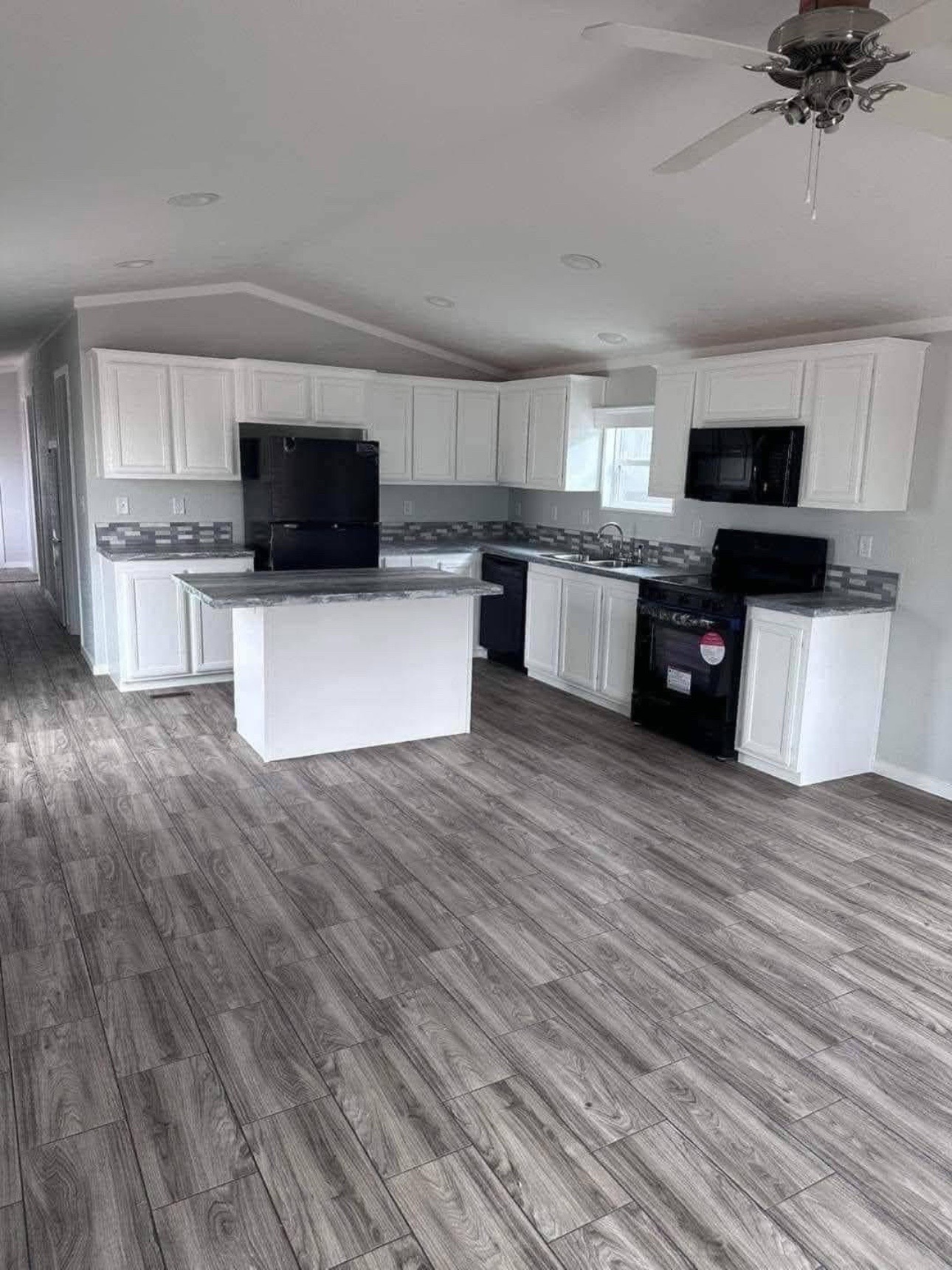 Modern kitchen with white cabinets, gray countertops, and matching tile backsplash. Black appliances contrast against the light wood floor. Bright and sleek.