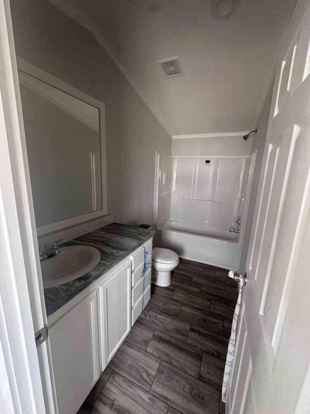 A small bathroom with gray tile flooring, white vanity with marble top, large mirror, white bathtub, and toilet. The space feels clean and modern.