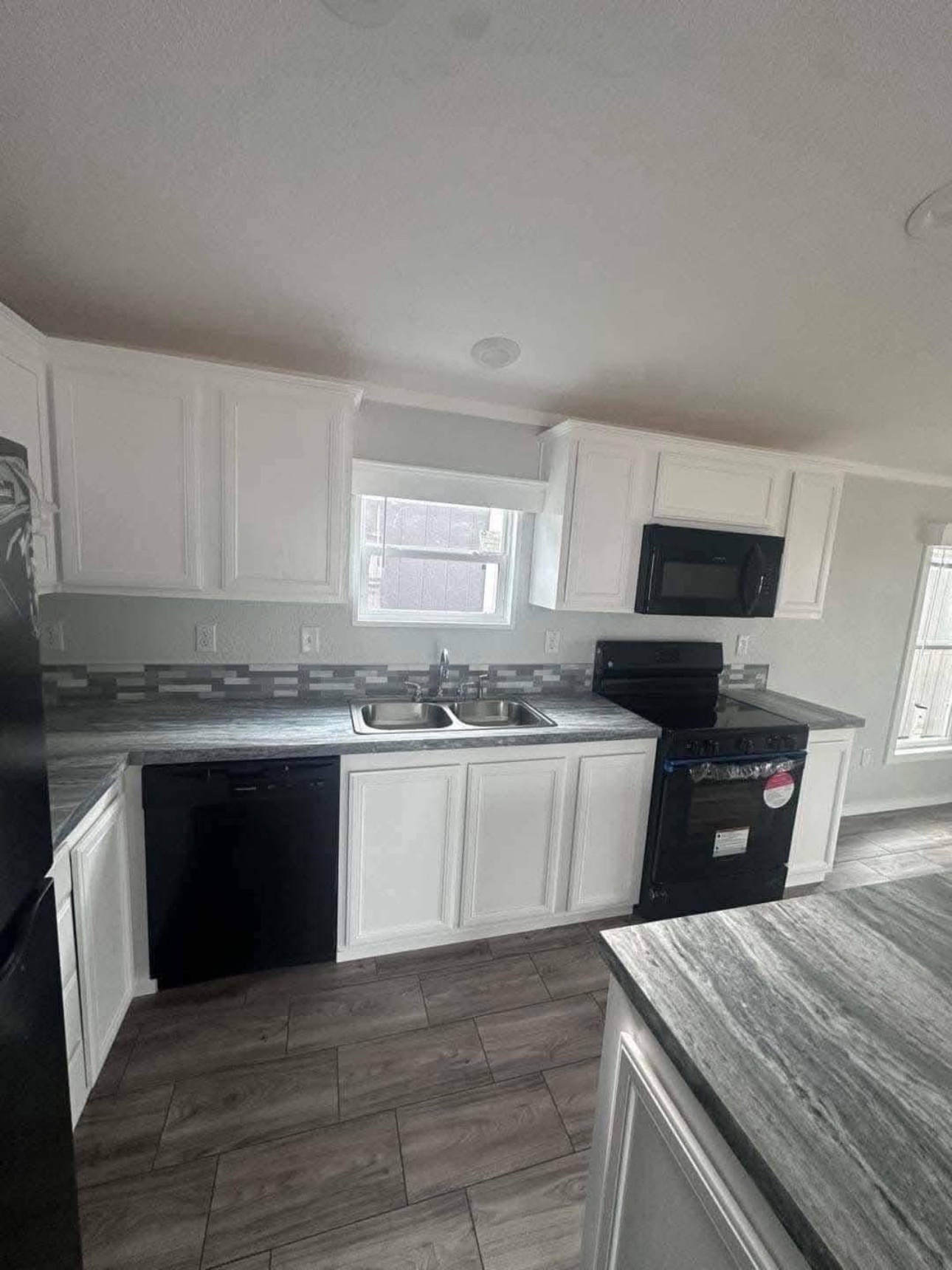 Modern kitchen with white cabinets, gray countertops, and tiled backsplash. Includes a black stove and microwave, stainless steel sink, and wood-look tiled floor.