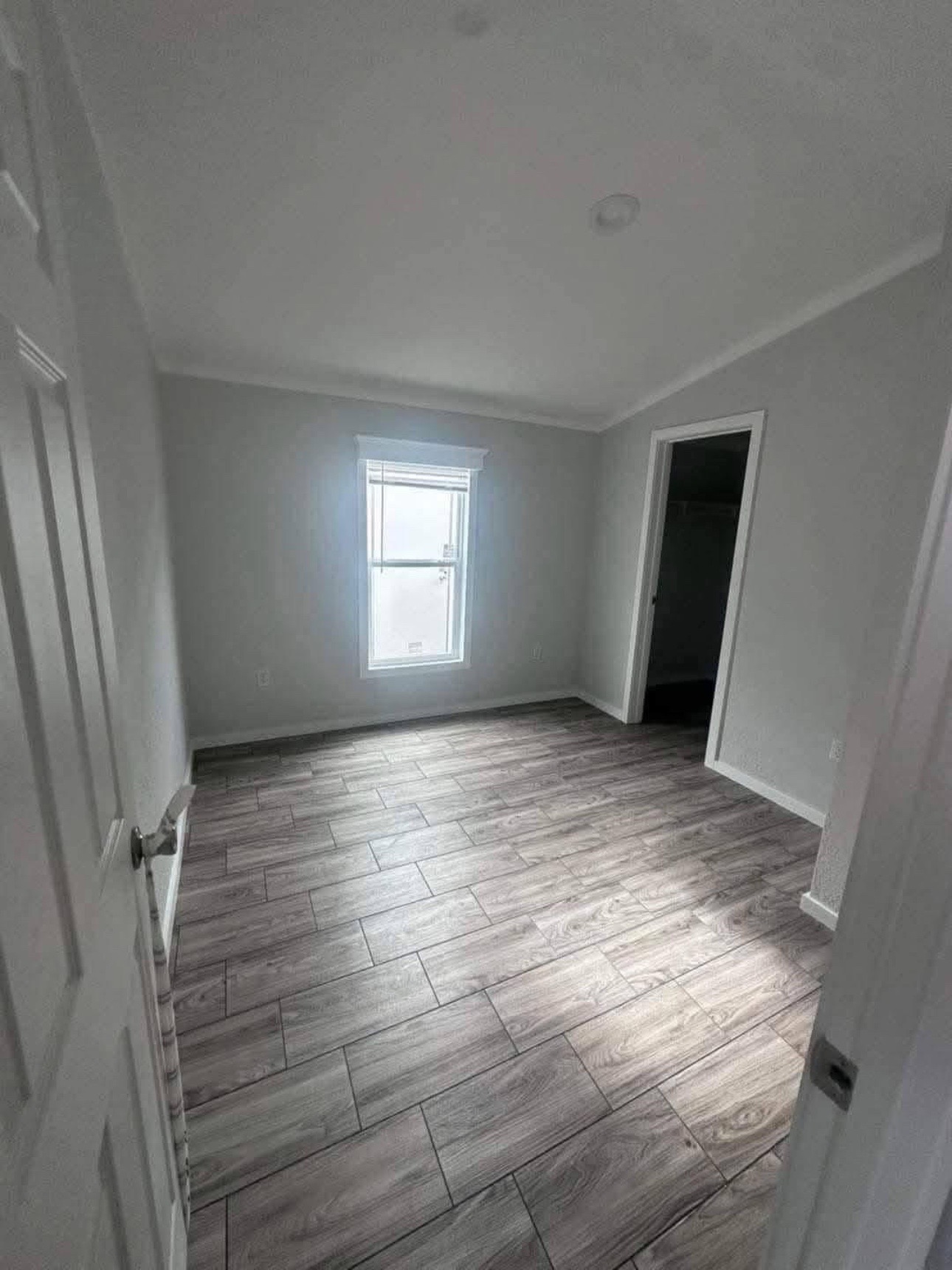 Small, empty room with light gray walls and wood-patterned tile floor. A window lets in soft light, and an open door reveals a dark closet. Minimalist and clean.