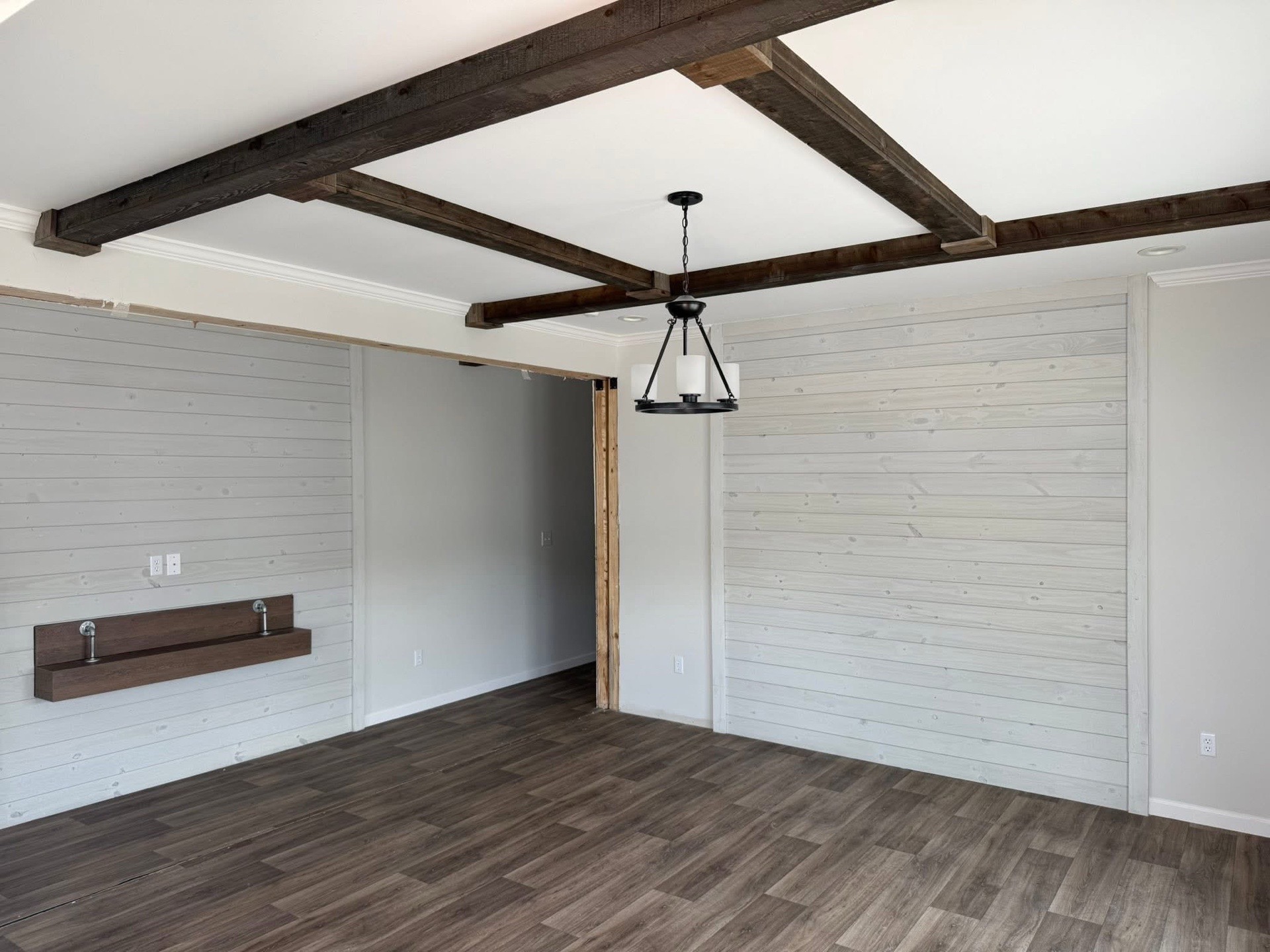 Spacious room with wood flooring and whitewashed plank walls. Dark wood ceiling beams and a farmhouse-style chandelier add rustic charm.