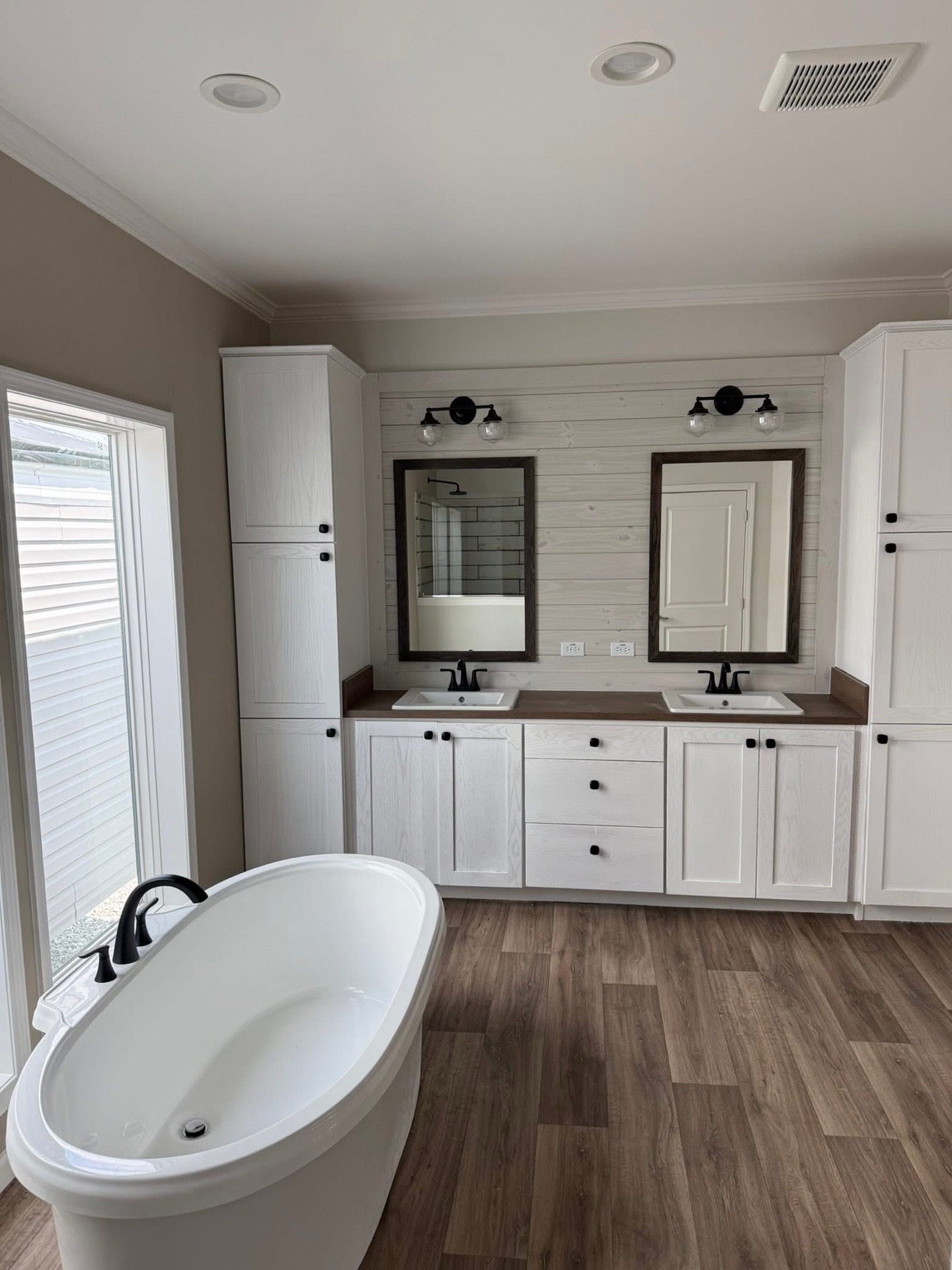 Stylish bathroom with a free-standing oval tub, dual sink vanity with white cabinetry, and black fixtures. Warm wood floors and soft lighting create a serene atmosphere.