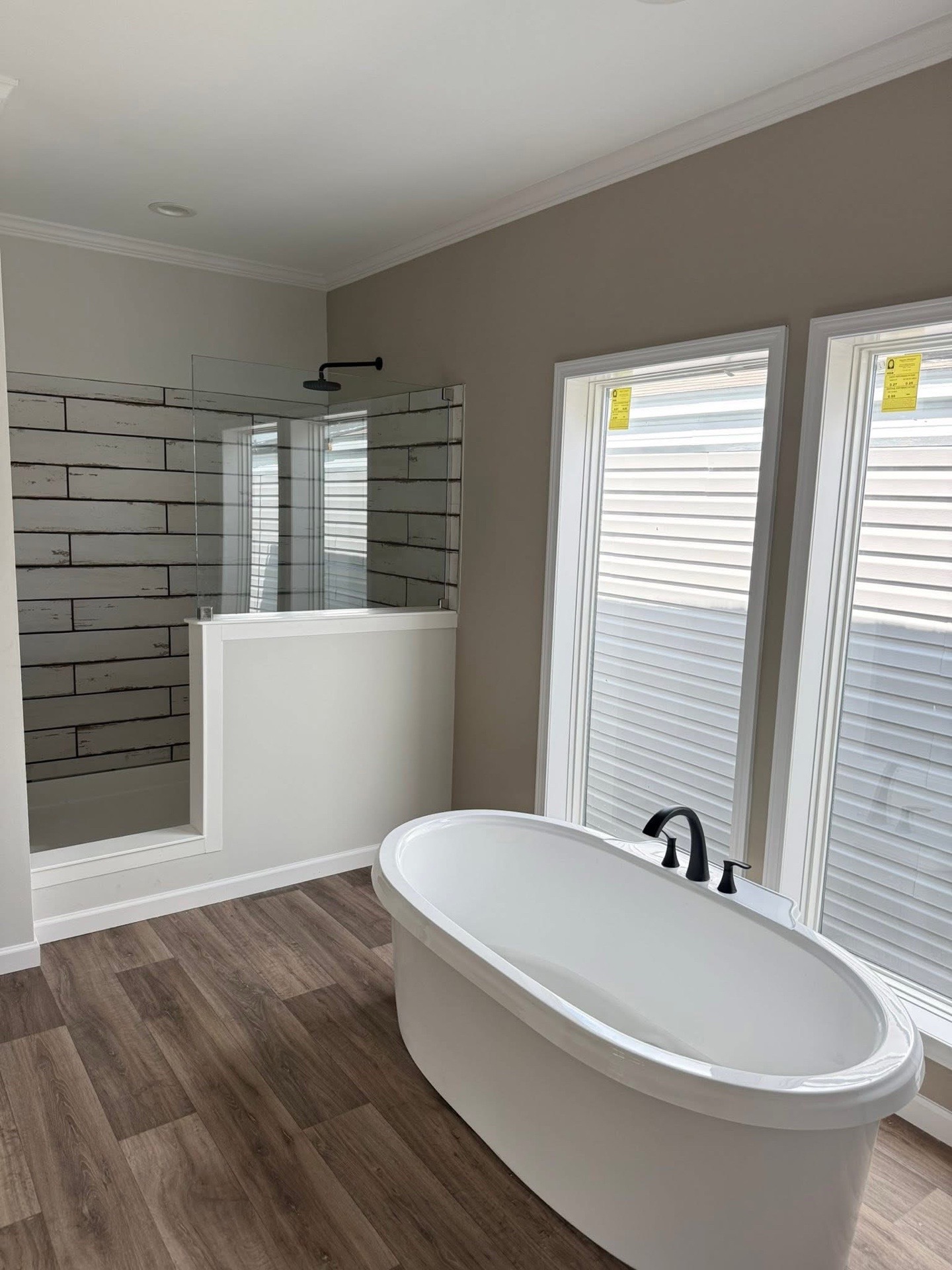 Modern bathroom with a free-standing white oval tub and black faucet, next to large windows. Shower with glass door and wood-patterned tiles. Warm, inviting.