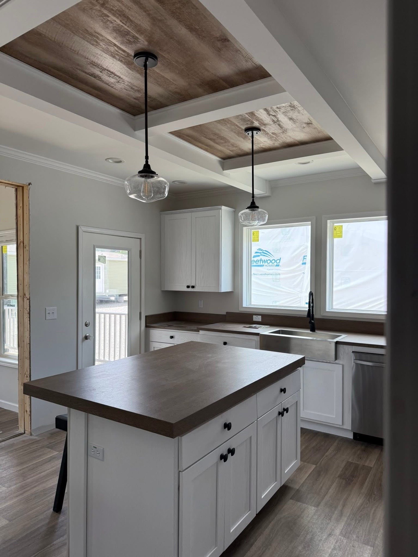Modern kitchen under construction with white cabinets, dark wood countertops, and island. Exposed beam ceiling with pendant lights; large windows, natural light.