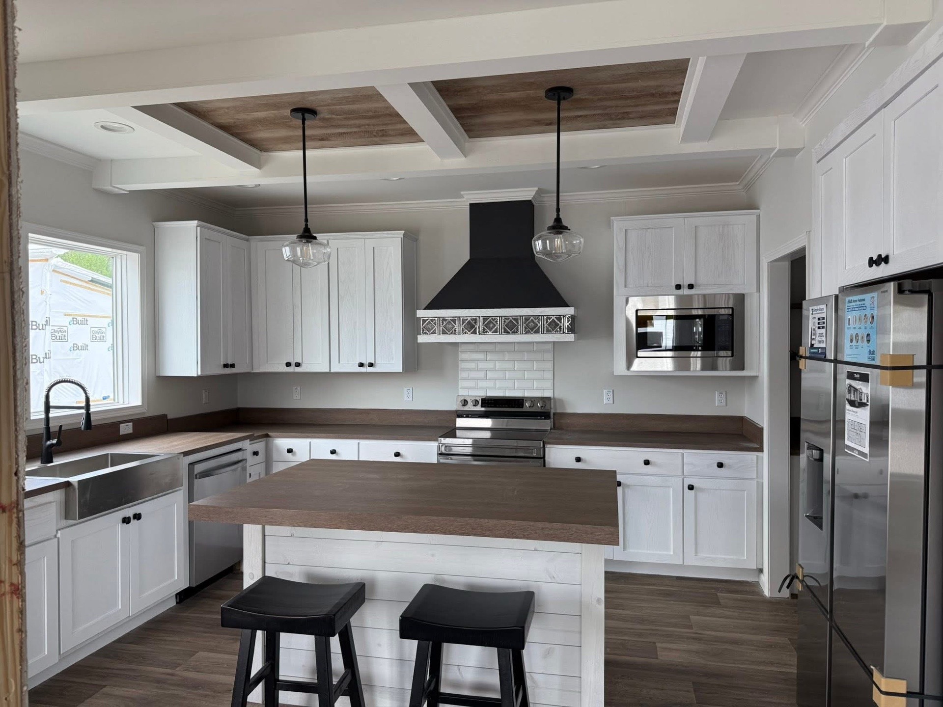 A modern kitchen with white cabinets, wood countertops, and a black range hood. It features a large island, pendant lights, and stainless steel appliances.