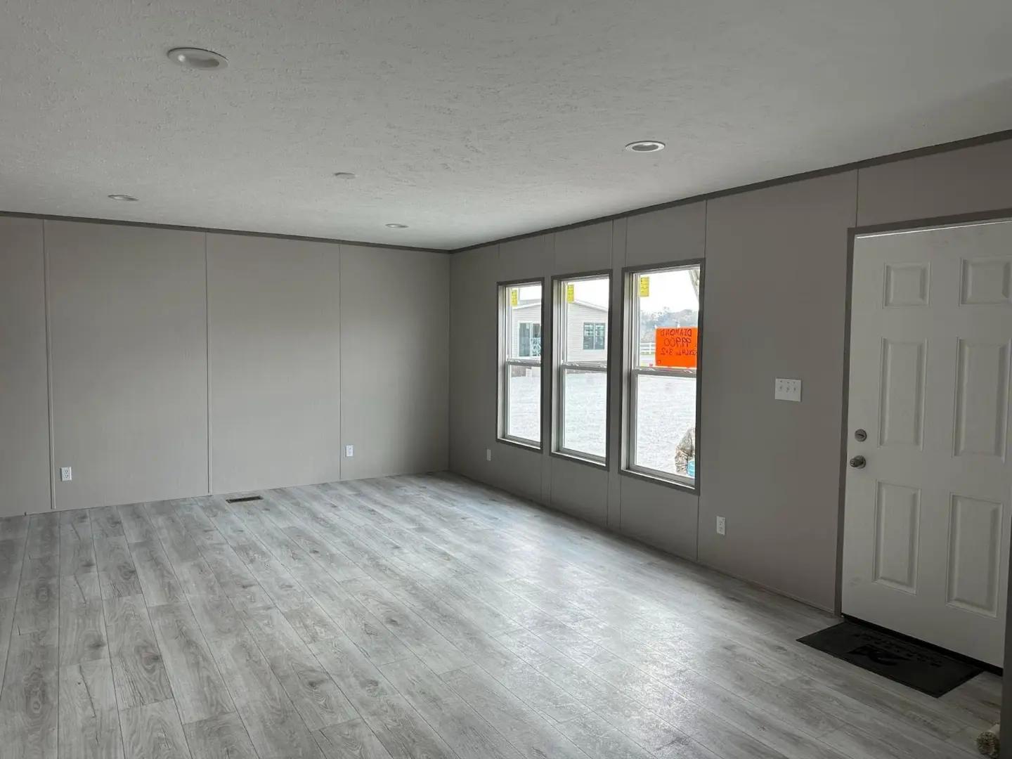 Empty room with light gray walls and wood flooring. Three large windows allow natural light, and a white door is on the right. Calm and neutral ambiance.