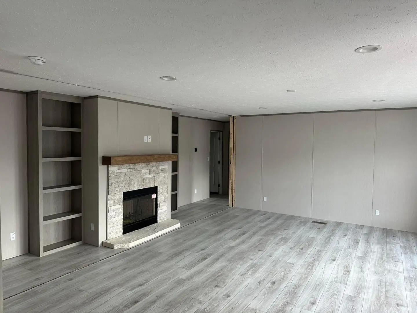 Spacious room with light gray walls and flooring. A stone fireplace with a wooden mantel is centered, flanked by built-in shelves. Calm, minimalist vibe.