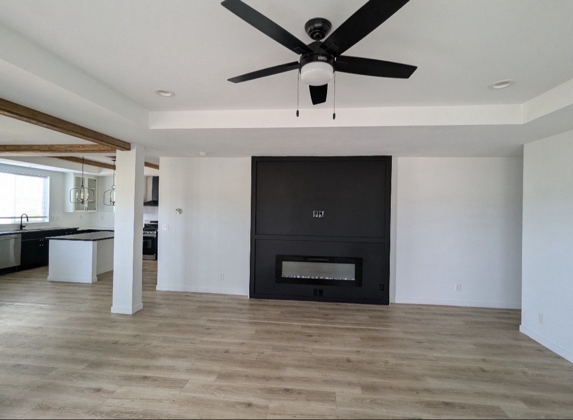 Spacious modern living room with light wood floors, a black accent wall featuring a fireplace, ceiling fan, and open layout to kitchen with pendant lights.