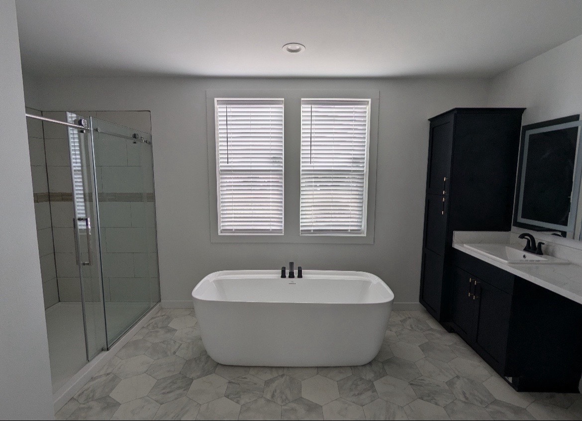 Modern bathroom with freestanding white tub, glass shower, and black vanity. Neutral tones, hexagon tile floor, and two windows create a serene atmosphere.