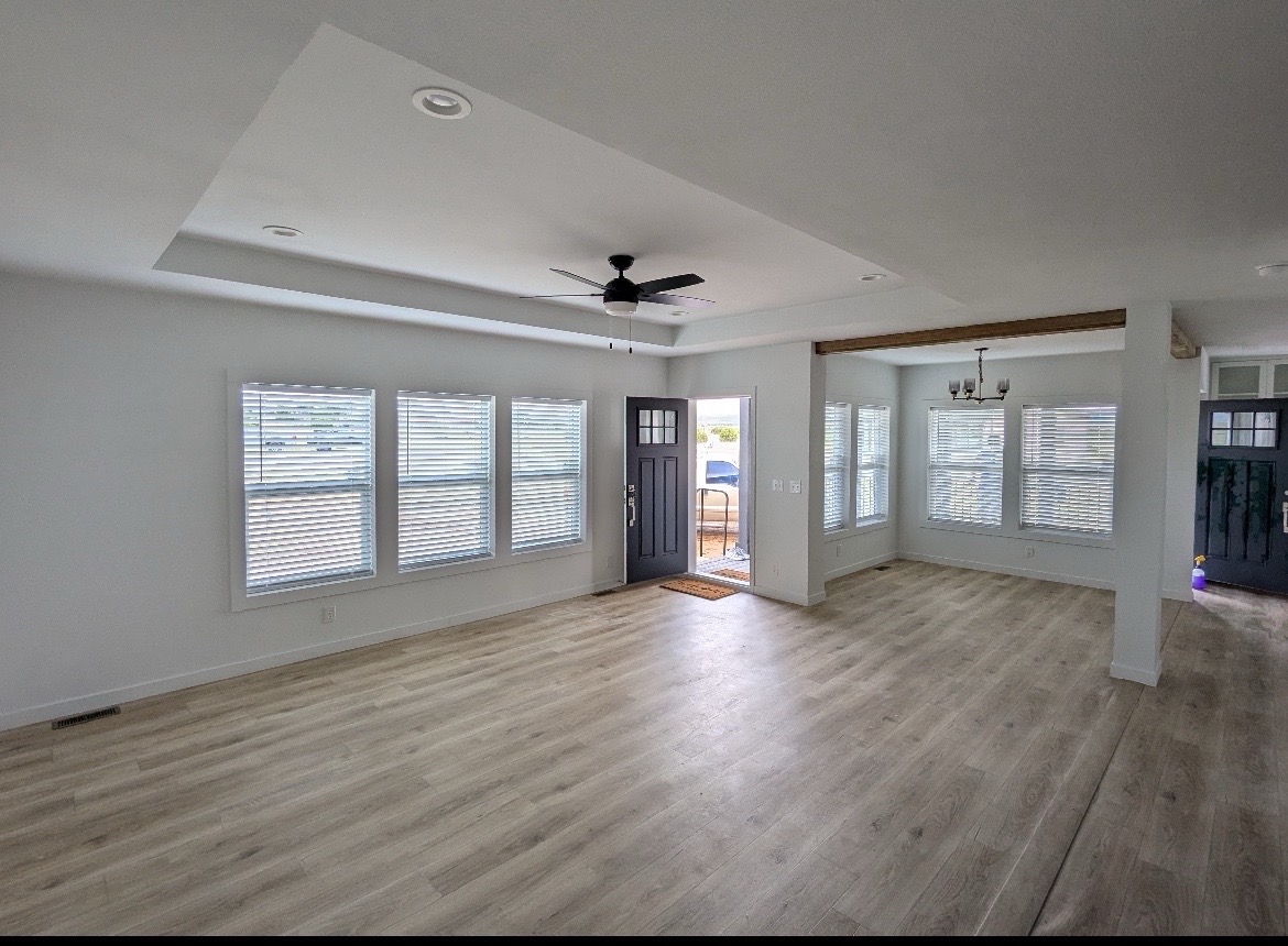 Spacious, bright living room with light wood flooring and white walls. Features large windows, gray front door, ceiling fan, and a chandelier.