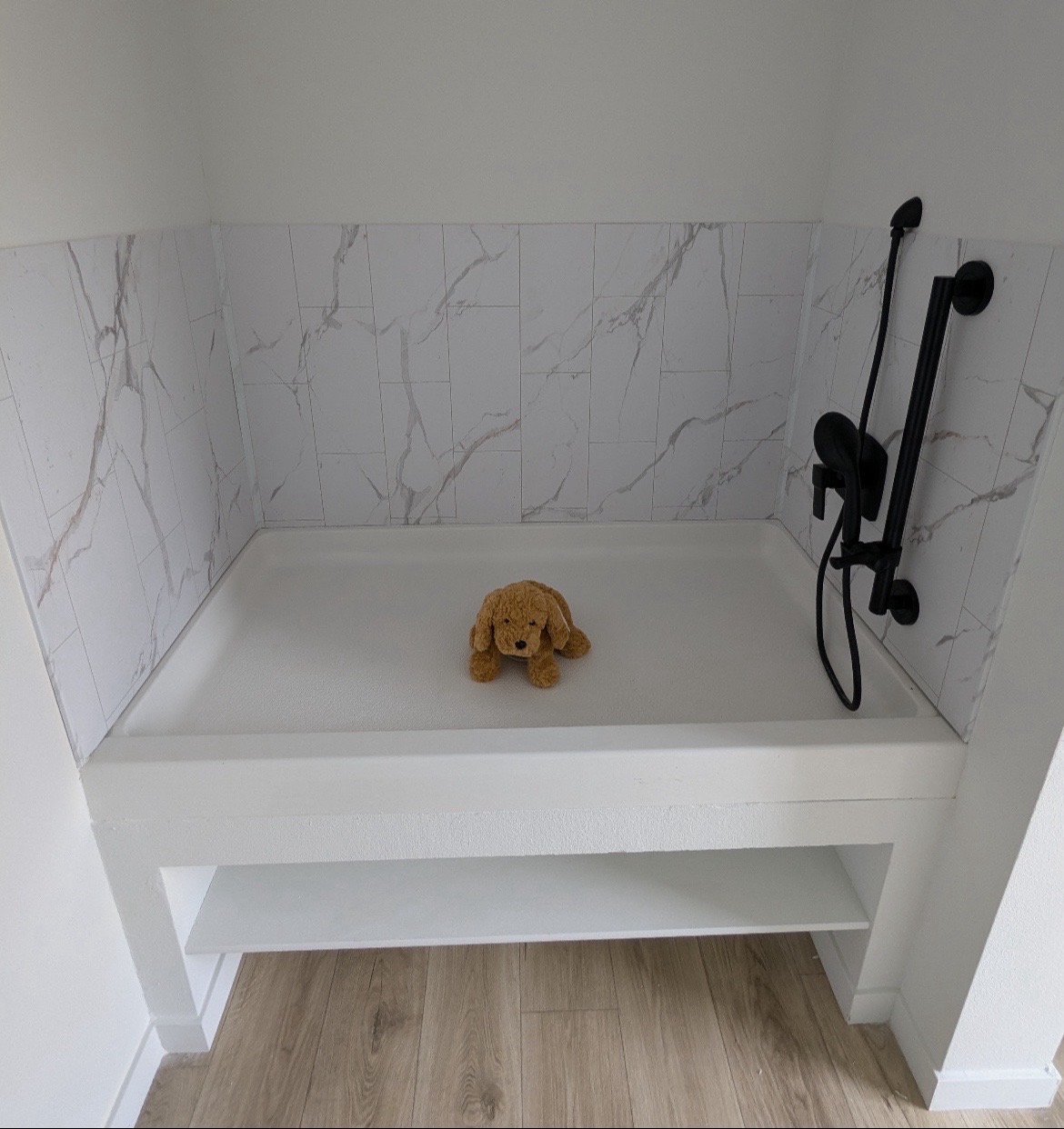 A small shower with white marble tiles and a sleek black showerhead. A plush brown dog toy sits on the basin, adding a cozy touch.