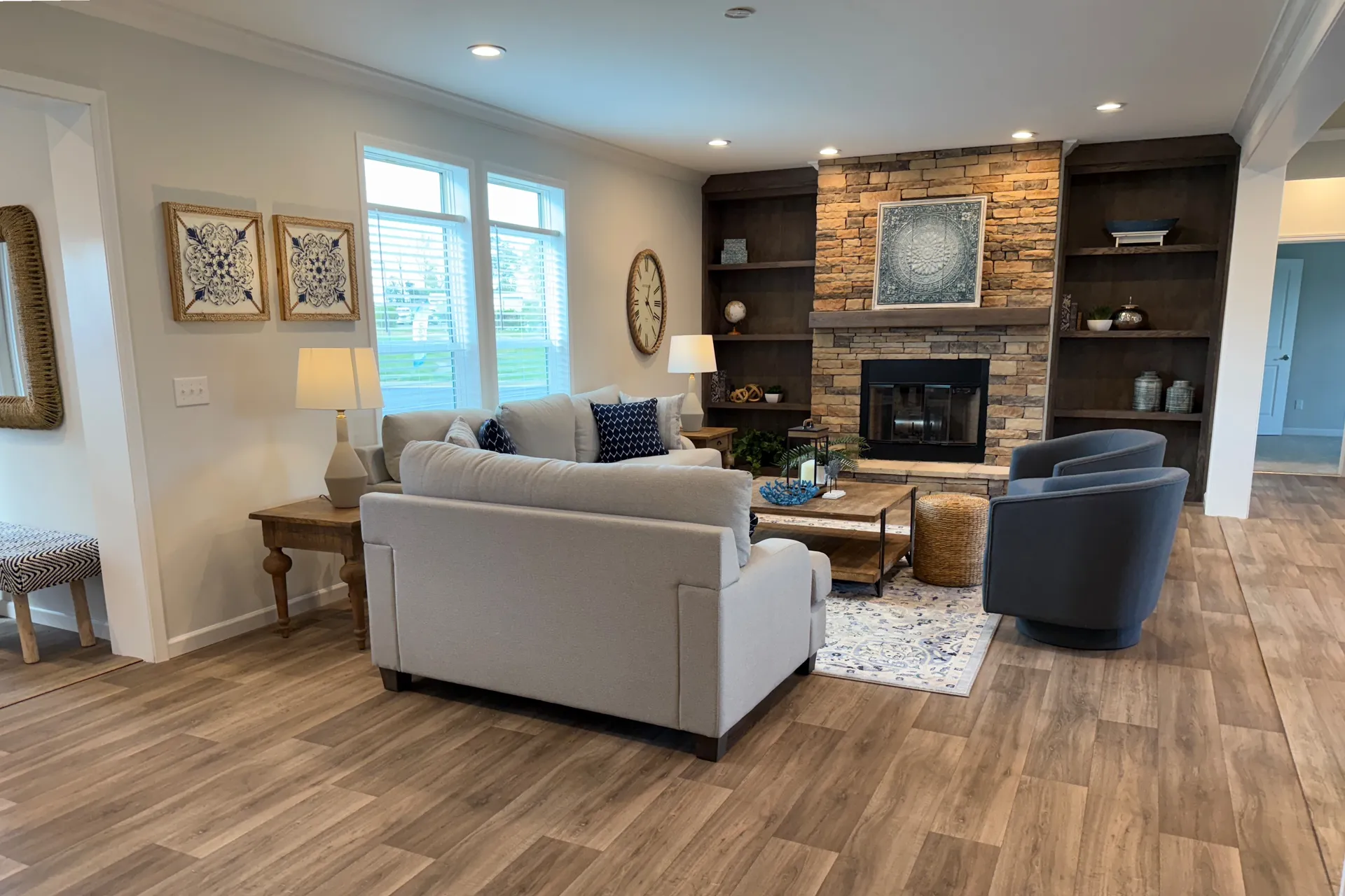 A cozy living room with a beige sofa, two gray chairs, and wooden coffee table. A stone fireplace flanked by shelves, soft lighting, and natural tones create a warm ambiance.