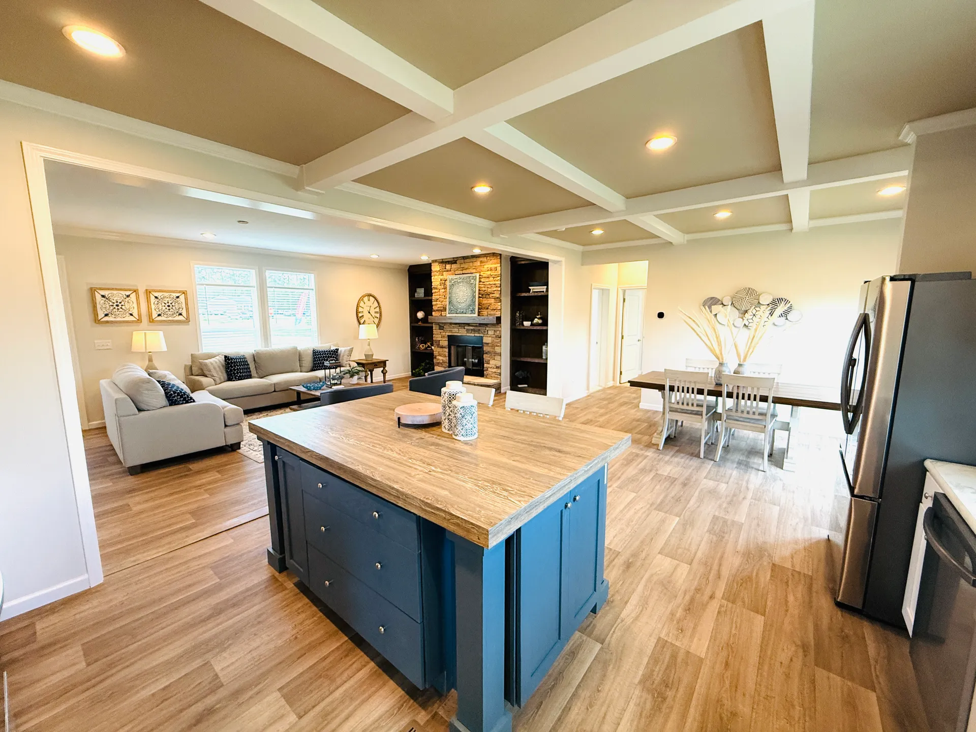 Spacious open-concept living room and kitchen with wooden floors, modern blue island, cozy gray sofas, dining table, and built-in fireplace. Warm, inviting ambiance.