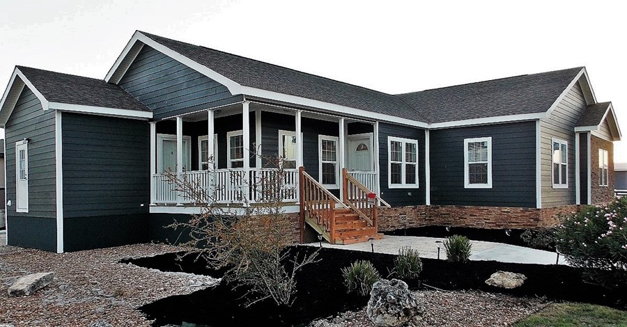 A modern, single-story house with dark gray siding and a wraparound porch set in a landscaped yard with shrubs and rocks, conveying a welcoming feel.