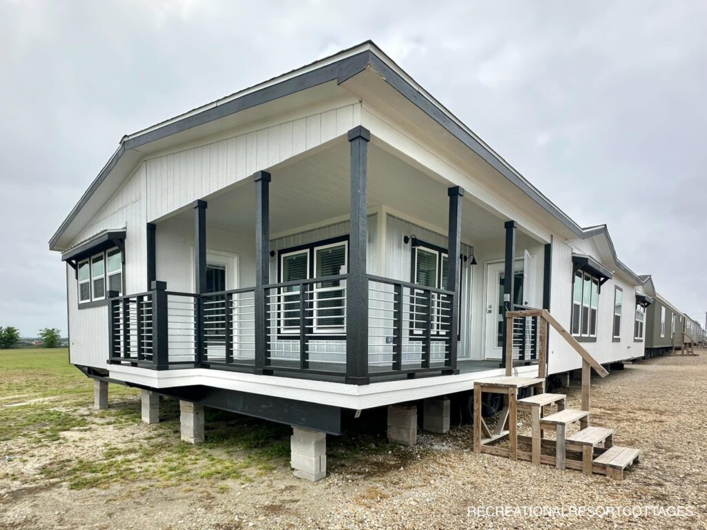A modern manufactured home with a white exterior and black trim. Features a wraparound porch with railings and steps. The sky is overcast.