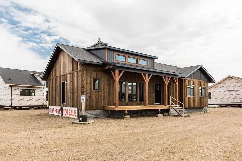 Wooden show home with large windows and welcoming porch on a dirt lot under a cloudy sky. "For Sale" signs are visible, suggesting availability.