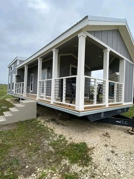 A modern tiny house on a trailer is set against a cloudy sky. It features a gray exterior, a spacious porch with white railing, and minimalist design elements.