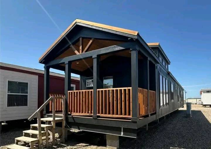 A modern, dark-colored tiny house on a gravel lot, featuring a wooden porch with railings, steps leading up, and a bright, clear sky overhead.