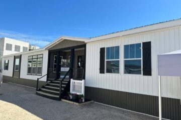 A modern, prefabricated building with white siding and black accents under a bright blue sky. The entrance features stairs and railings, creating a welcoming appearance.
