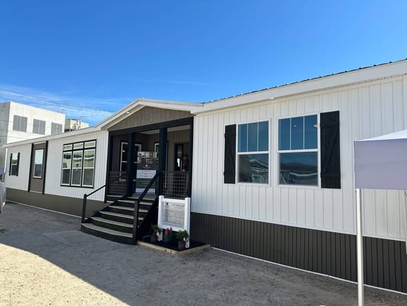 A modern, prefabricated building with white siding and black accents under a bright blue sky. The entrance features stairs and railings, creating a welcoming appearance.