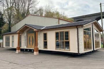 A modular home with large windows and wooden accents, situated on a concrete lot. It's backed by tall trees and a viaduct, under a cloudy sky.