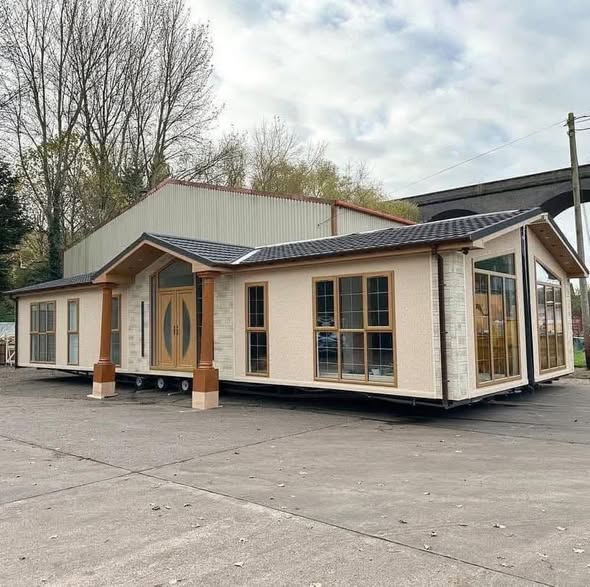 A modular home with large windows and wooden accents, situated on a concrete lot. It's backed by tall trees and a viaduct, under a cloudy sky.