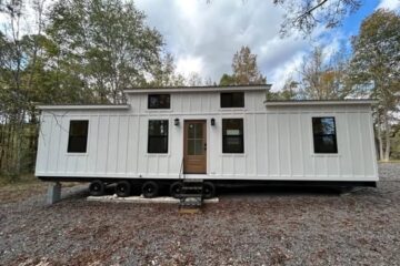 White modular tiny house with black-framed windows, set on wheels, on a gravel lot. Surrounded by trees, under a partly cloudy sky. Peaceful ambiance.