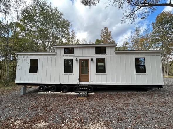 White modular tiny house with black-framed windows, set on wheels, on a gravel lot. Surrounded by trees, under a partly cloudy sky. Peaceful ambiance.