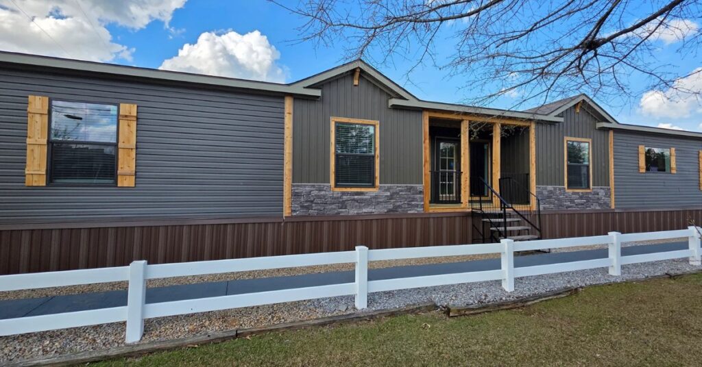 Single-story home with gray siding, wooden accents, and stone trim. Front door has steps; white fence and grass lawn in foreground. Sky is blue.