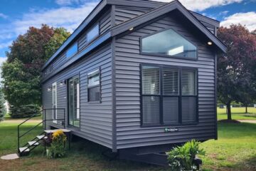 A modern gray tiny house on a grassy field with large windows and a sloped roof. Potted plants and flowers accent the entrance. Sky is partly cloudy.