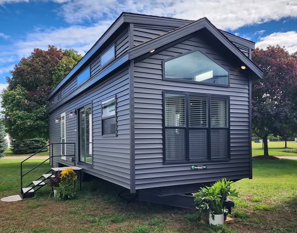 A modern gray tiny house on a grassy field with large windows and a sloped roof. Potted plants and flowers accent the entrance. Sky is partly cloudy.